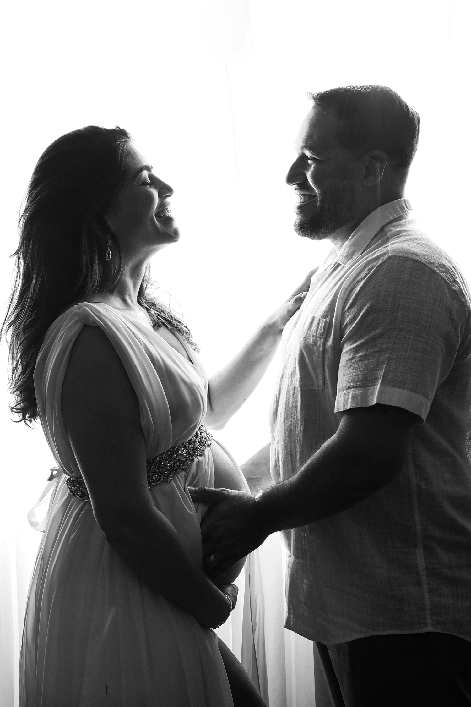 Fotografia em preto e branco de um casal durante ensaio de gestante em estúdio. A mulher veste um vestido fluido longo e o homem está com a mão carinhosamente apoiada na barriga dela. Eles sorriem um para o outro, iluminados por uma luz de fundo que destaca a silhueta de ambos.