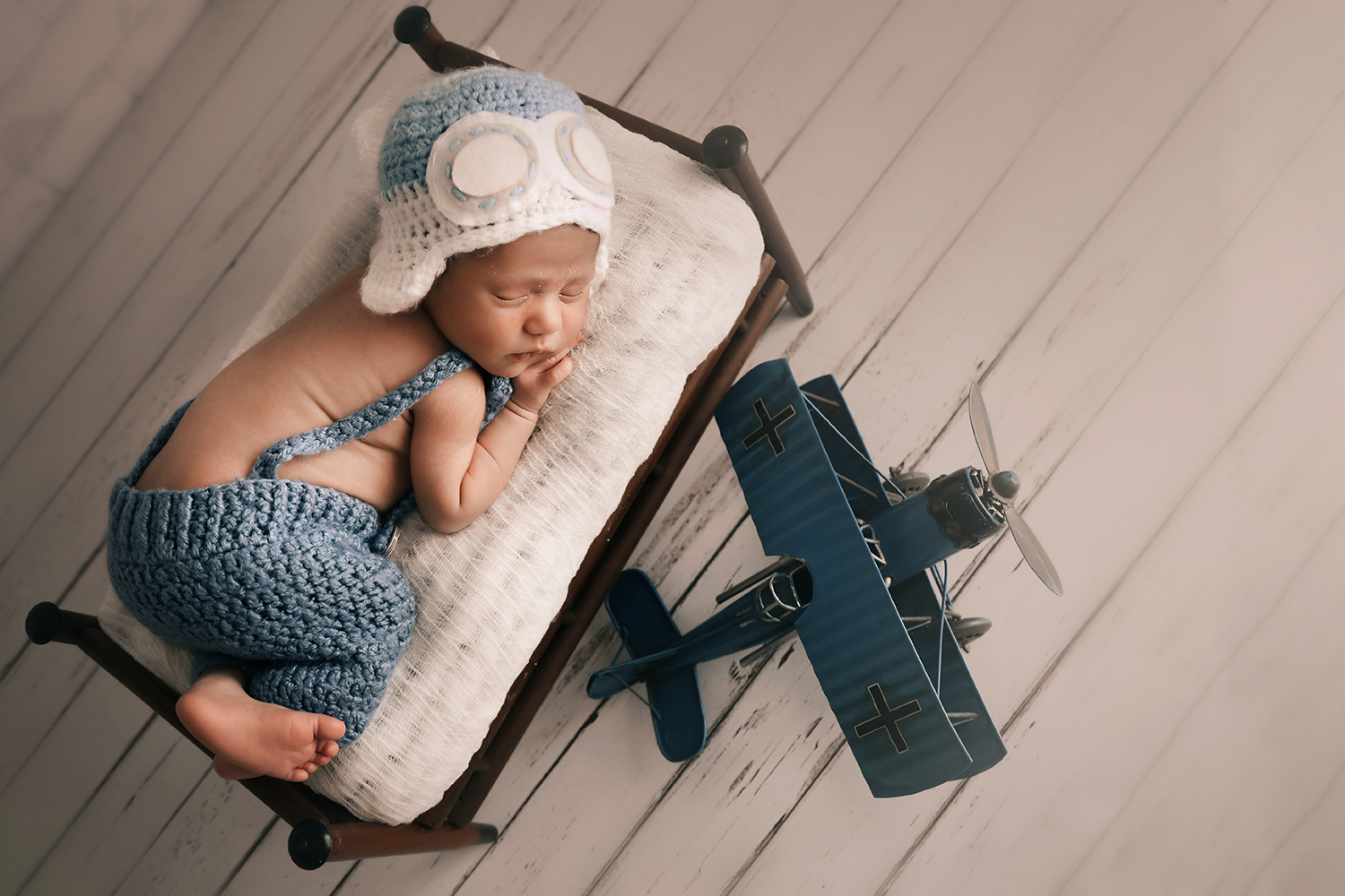 Bebê recém-nascido dormindo de lado em uma caminha de madeira em um estúdio fotográfico na Zona Leste de São Paulo. O bebê usa uma touca de crochê de aviador com óculos e calça azul. Ao lado da cama há um pequeno avião de brinquedo azul vintage sobre um fundo de piso de madeira clara.