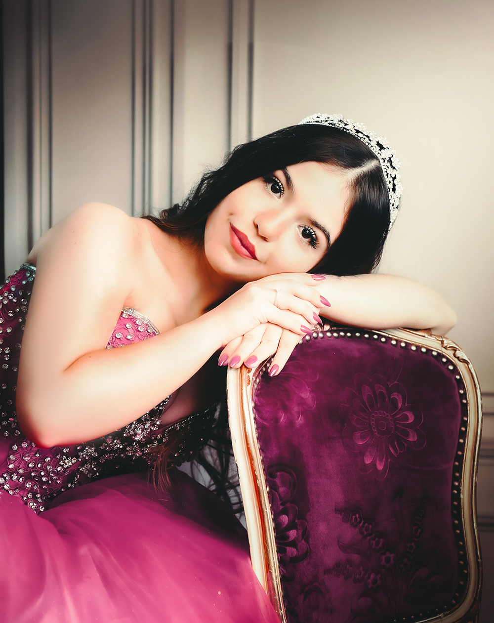 Debutante vestindo vestido de gala rosa e tiara, sorrindo e apoiada numa cadeira de veludo em ensaio de 15 anos no Estúdio Roni Sanches.