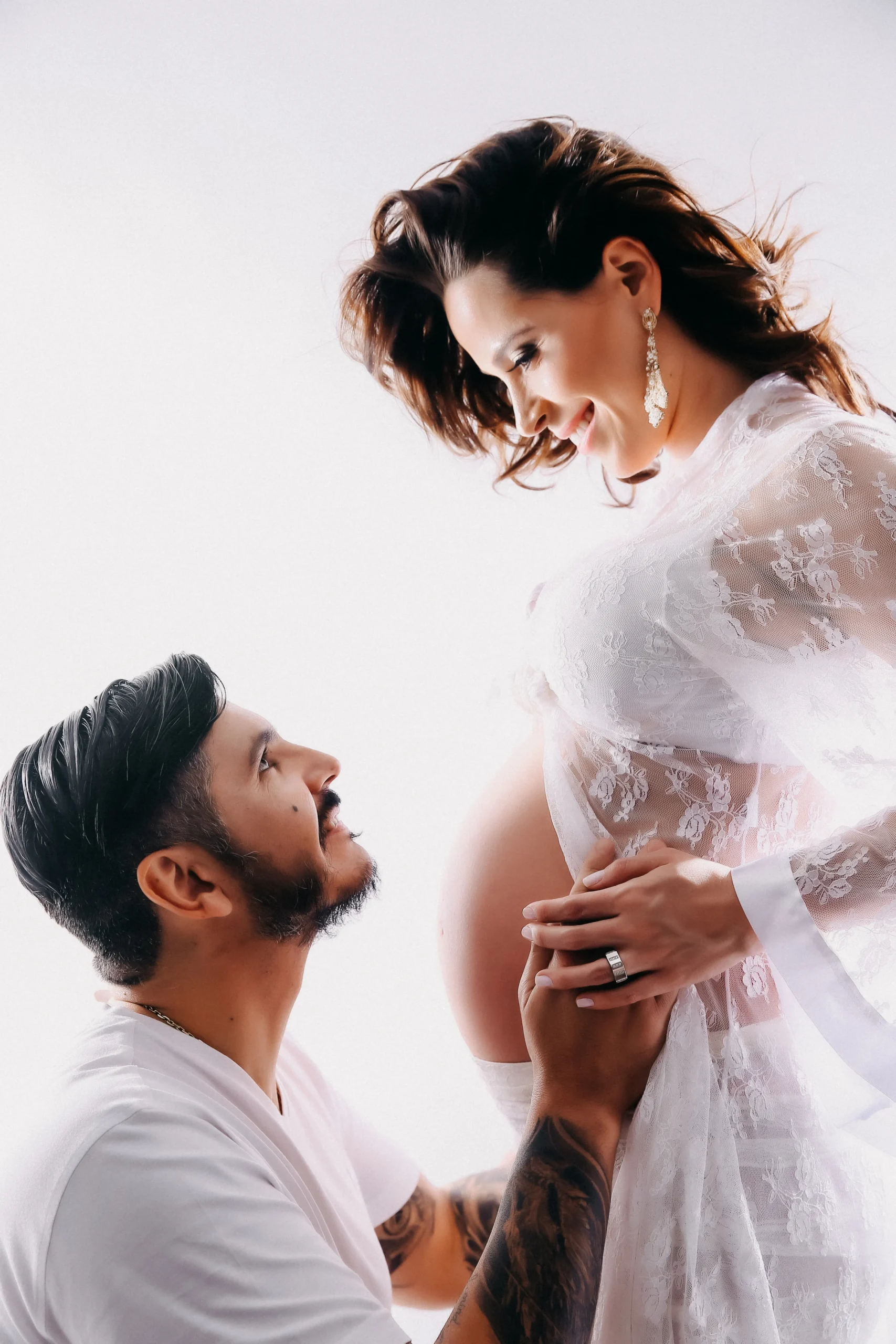 Fotografia em estúdio com fundo claro e bem iluminado de um casal. A mulher grávida está em pé vestindo um delicado vestido longo de renda branca. O parceiro está posicionado mais abaixo, com as mãos na barriga de gestante dela. Eles se olham sorrindo com amor e cumplicidade.