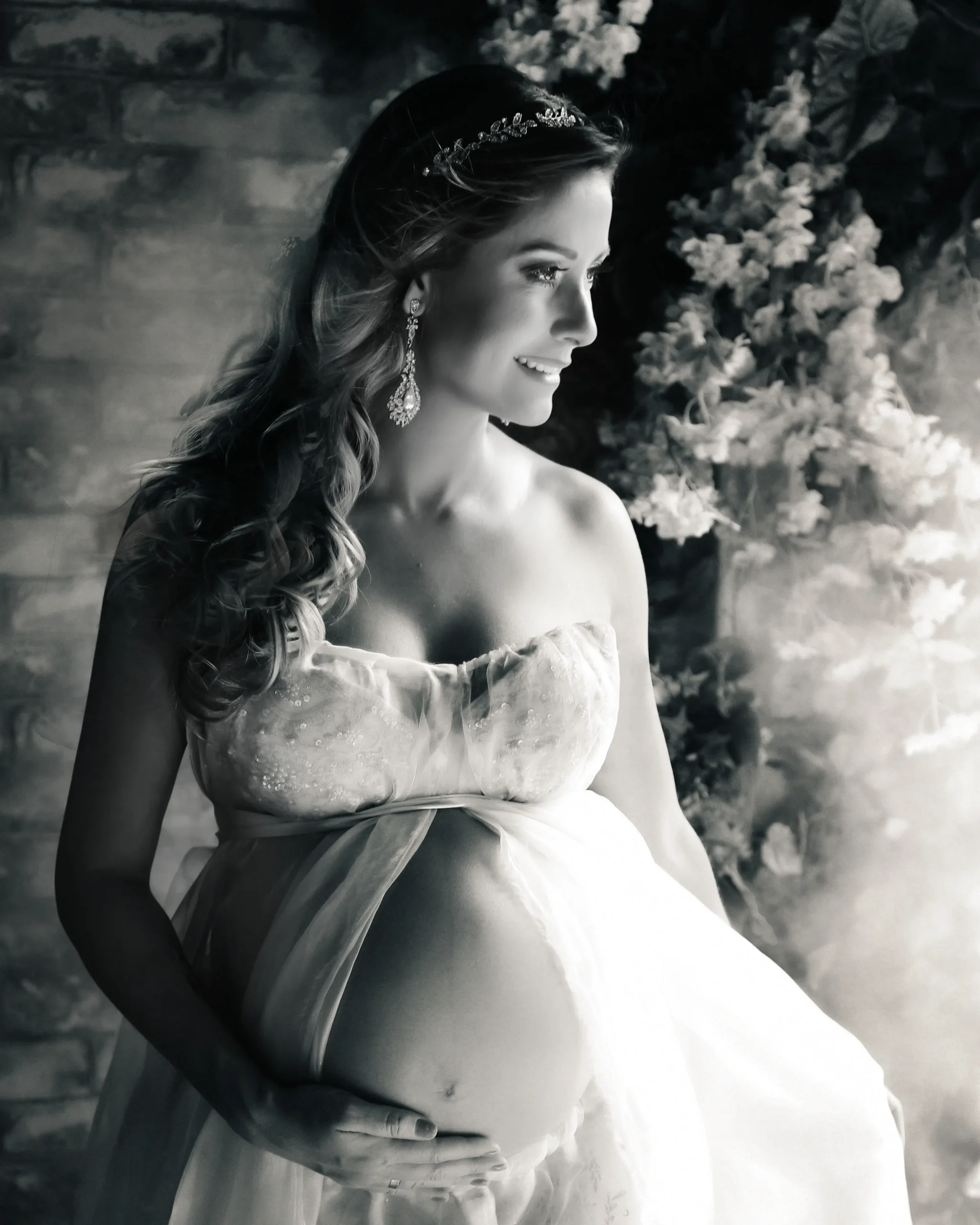 Fotografia Fine Art em preto e branco de uma mulher grávida sorrindo suavemente. Ela tem cabelos longos com uma tiara delicada e brincos. Veste um top e uma saia fluida que deixam sua barriga de gestante à mostra. Ela está segurando a barriga com carinho. Atrás dela há um cenário elegante com um grande arranjo de flores claras desfocadas.