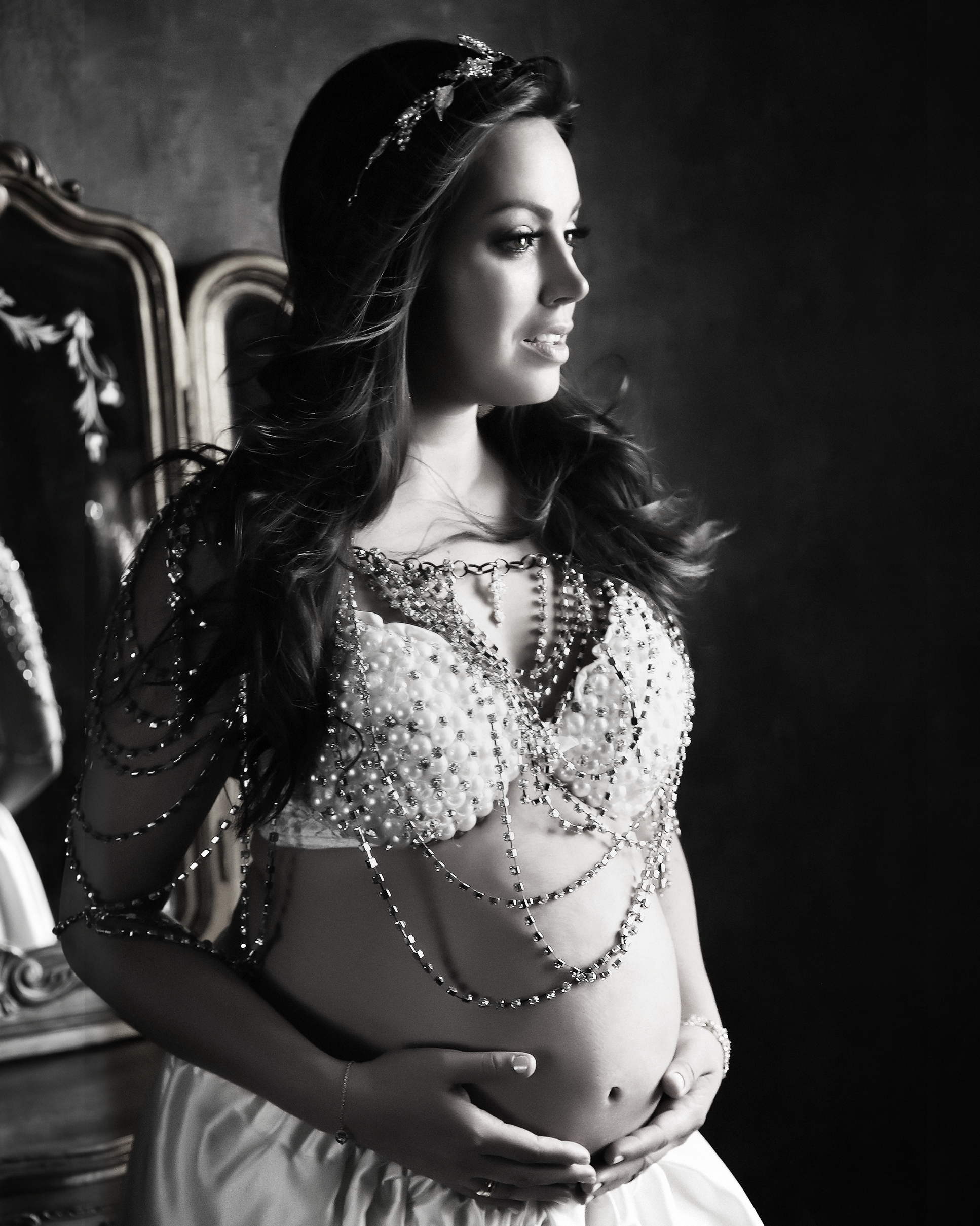 Fotografia em preto e branco de uma mulher grávida em estúdio. Ela está de perfil, olhando para frente e segurando a barriga. Veste um top ornamentado com correntes e pérolas, saia longa e uma tiara brilhante no cabelo escuro ondulado, com um fundo de estúdio texturizado.