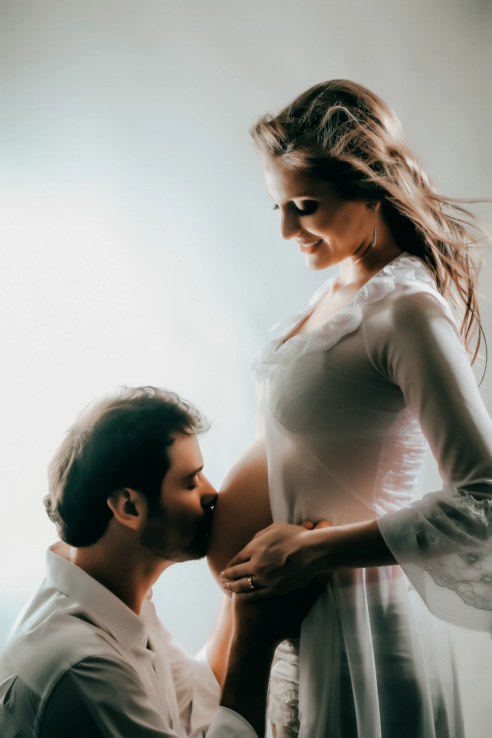 Fotografia de estúdio de um casal esperando bebê. O homem está agachado beijando a barriga da mulher grávida. Ela usa um vestido branco fluido e sorri afetuosamente olhando para ele. Uma luz suave ilumina o contorno do casal por trás, criando uma atmosfera romântica e emocionante.