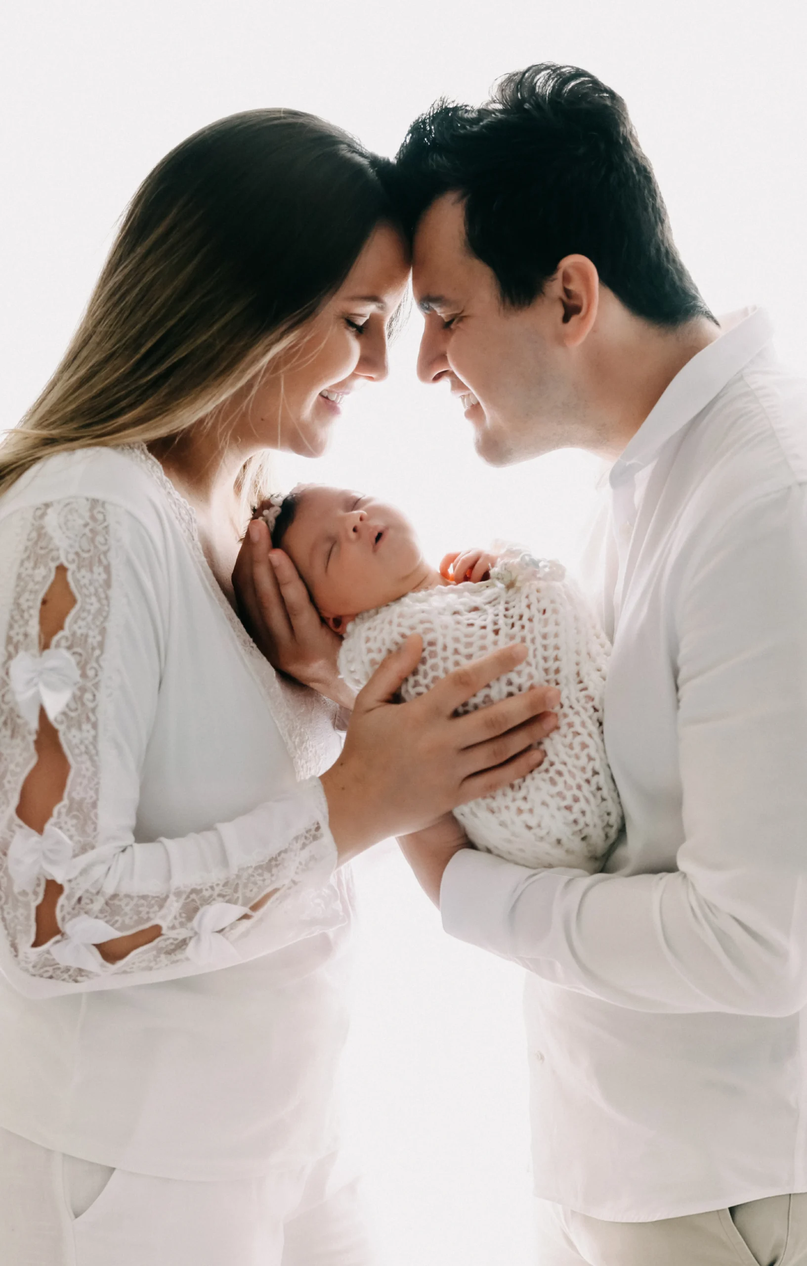 Fotografia em fundo branco e luminoso de um casal segurando seu bebê recém-nascido. A mãe e o pai, vestindo roupas brancas e leves, tocam as testas carinhosamente e sorriem enquanto olham para o bebê deitado nas mãos do pai. O bebê dorme sereno, enrolado em uma manta de tricô macia e branca.