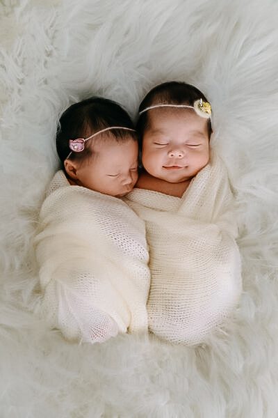 Fotografia vista de cima de dois bebês recém-nascidos gêmeos dormindo profundamente e sorrindo de forma suave. Eles estão enrolados lado a lado em tecidos brancos texturizados (swaddle) e usam tiaras finas com pequenos detalhes florais na cabeça. Estão deitados sobre um fundo de pelúcia branca muito macia.