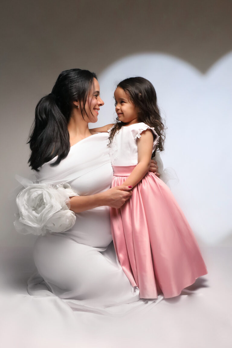 Ensaio de gestante em São Paulo com mãe e filha de vestidinho rosa sorrindo juntas em estúdio. Fotos de grávida repletas de amor e cumplicidade. Book gestante SP.
