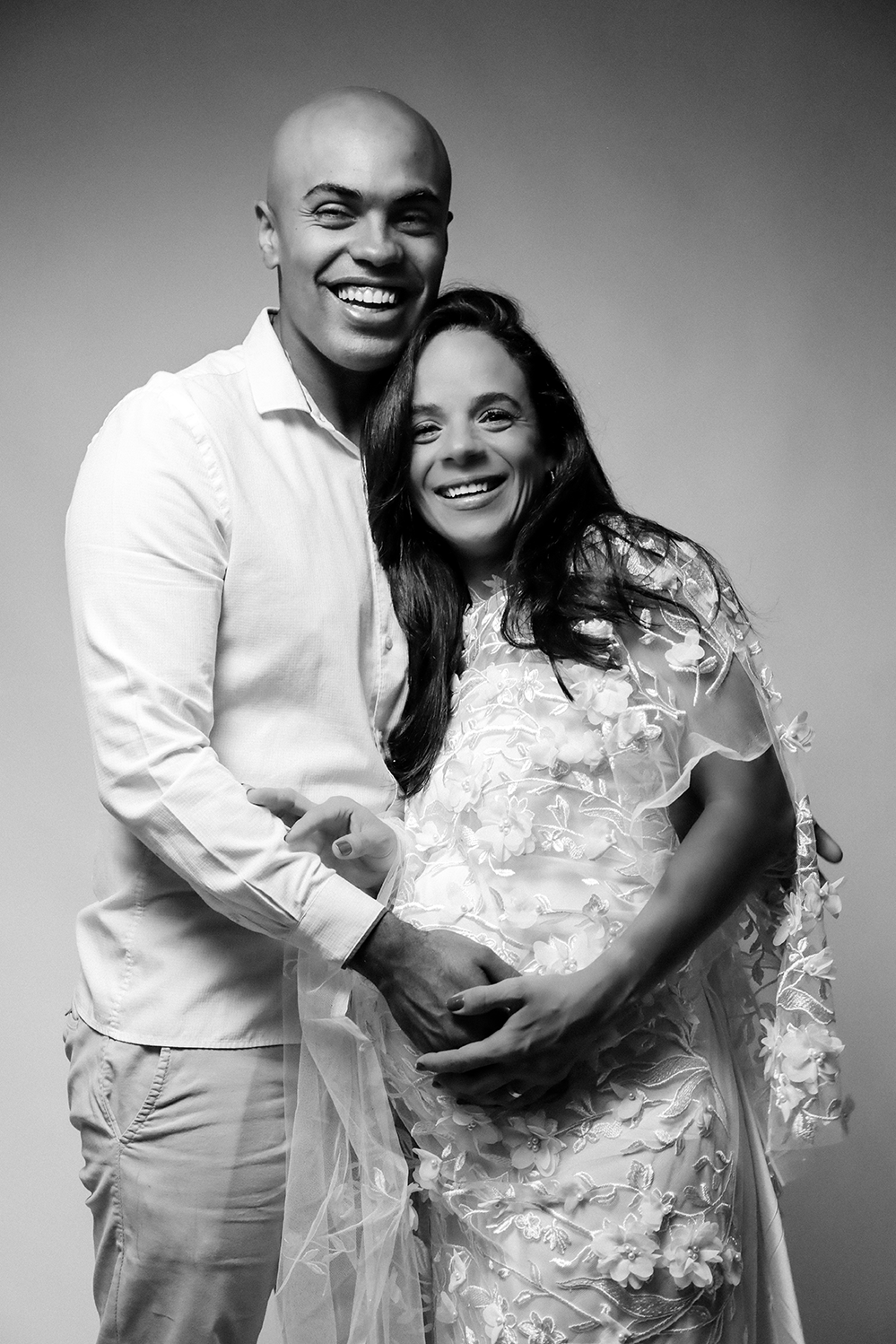 Ensaio gestante casal em preto e branco em SP: gestante de vestido rendado e marido sorridentes segurando a barriga em estúdio.